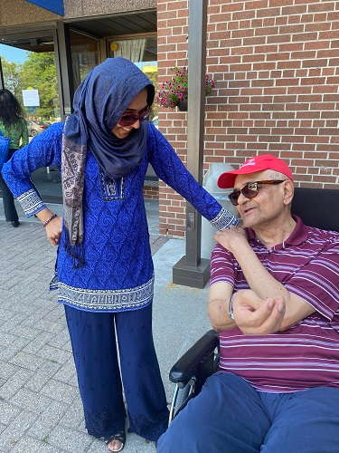 10 Little Ways to Show Love to an Elderly Parent Every Day, Nargis smiling while standing beside her father, who is seated in a wheelchair, engaging in a warm conversation outside a brick building.